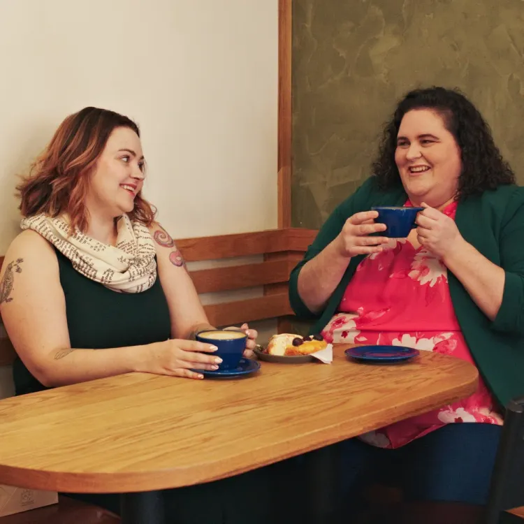 A photo of Robyn and Alicia in a cafe, sitting next to and looking at eachother, holding cups of coffee.