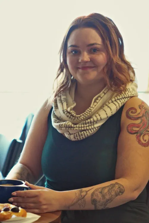 A portrait of Robyn, wearing a green shirt and a scarf, holding a cup of coffee.